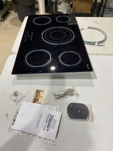 Black five-burner ceramic glass cooktop with mounting hardware and manual on white surface